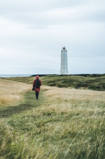 woman in red dress walks on green grass leading to white lighthouse