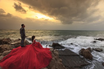 Woman in Red Gown and Man in Gray Suit Standing and Sitting on Boulders in Front of Ocean