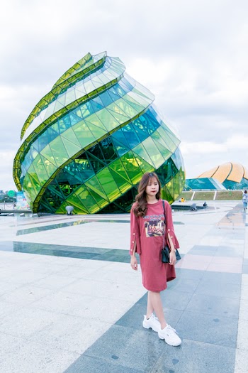Woman in Red Long-sleeved Top Wearing White Sneakers Walking in Front of Green and Blue Glass Building