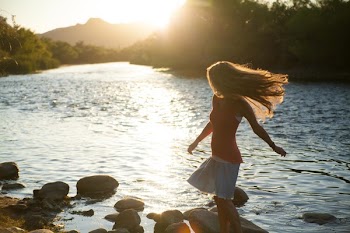 Woman in Red Tank Top Standing on Stone Near Body of Water during Daytime