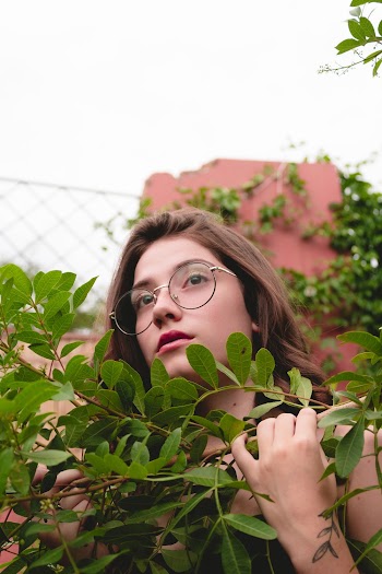 Woman in Silver-framed Clear Eyeglasses Standing Near Fence Holding Green Plant Duting Daytime