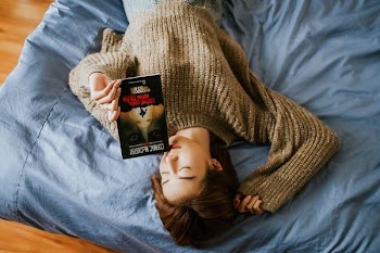 woman in sweater holding book while lying on bed
