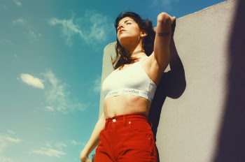Woman in White Calvin Klein Sports Bra and Red Bottoms Leaning on Gray Wall Under Gray Sky