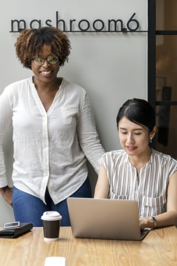 Woman in White Dress Shirt Standing Beside Woman Using Laptop