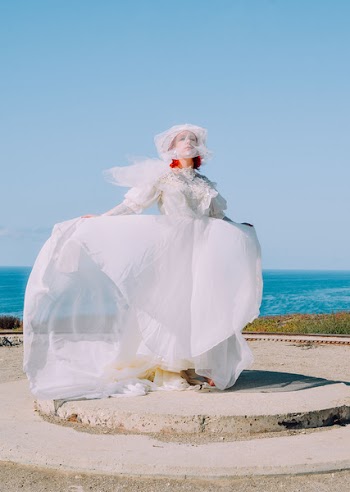 Woman in White Lace Wedding Dress Standing on Gray Pavement Near Body of Water during Daytime