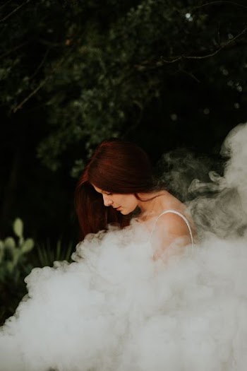 woman in white spaghetti strap sleeveless shirt standing near trees covered her body of smoke