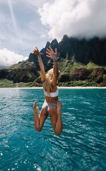 woman jumping on cliff into water while raising hands and folding both knees