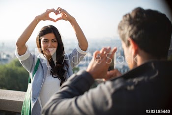 Woman posing with heart-shape hands for boyfriend on sunny balcony