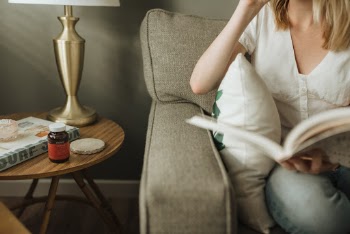Woman reading book on living room couch in the morning. Side table holds magazines, a lamp, and a Nouri Gut Rest probiotic bottle.
