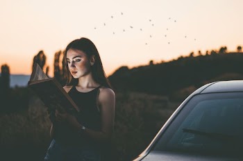 woman reading book outdoor beside car during dawn