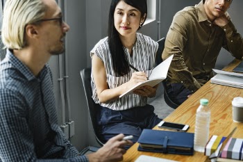 Woman Sitting Down Near Two Men Holding Up Pen and Notebook