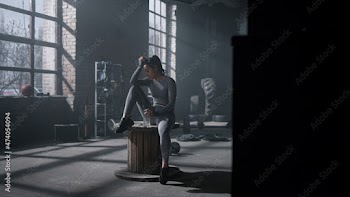 Woman sitting on wooden box in gym. Sportswoman holding sports bottle in hand
