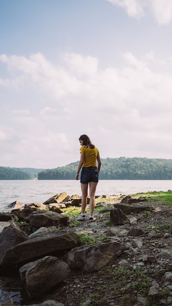 Woman Standing Near Rocks Facing Body of Water Under White Sky