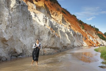 Woman Standing on Body of Water Near Rocky Hill Under Blue and White Skies
