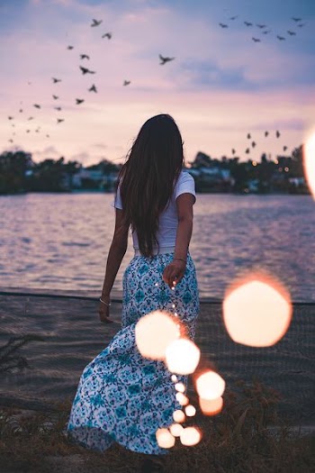 woman standing on green grass field in front body of water under flock of birds