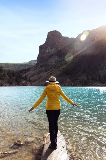 Woman Standing On Log Facing Body Of Water And Mountains 