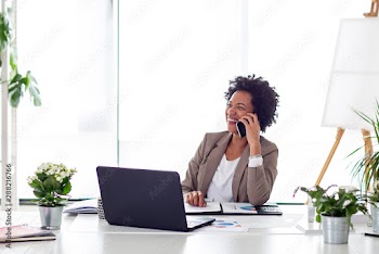Woman using mobile phone while sitting at the desk in office with laptop in front of her