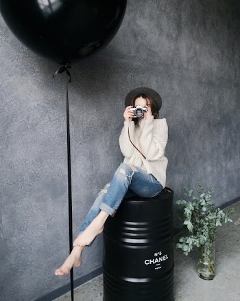 Woman Using Silver Camera Sitting on Black Container Drum