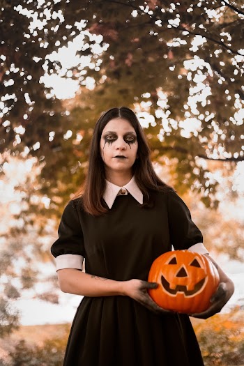 Woman Wearing Black and White Collared Dress With Halloween Makeup Holding Jack-o'-lantern Near Green Leaf Tree