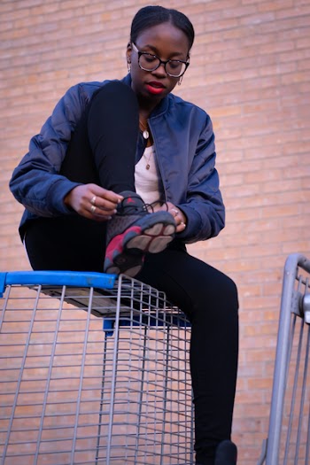 Woman Wearing Black Bomber Jacket and Air Jordan 13 Shoes Sitting on Gray Wire Crate