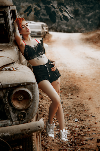 Woman Wearing Black Bra And Black Shorts Leaning On Wrecked Vehicle Beside Dirt Road