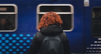 woman wearing black hooded jacket and black backpack standing in front of moving blue train