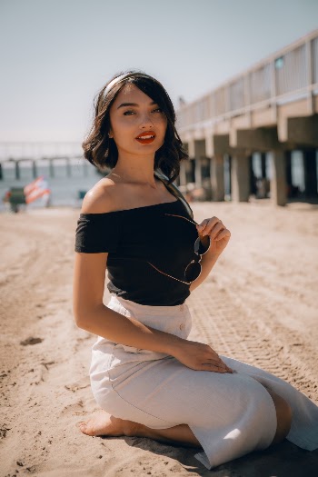 Woman Wearing Black Off-shoulder Top and White Skirt Sitting on Sand While Holding Sunglasses