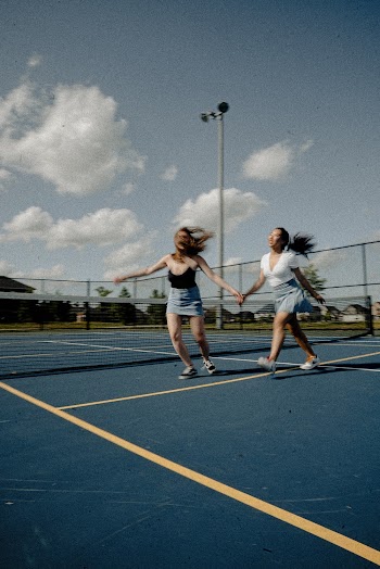 Woman Wearing Black String Strap Top on Tennis Court Besides Woman Wearing White Top