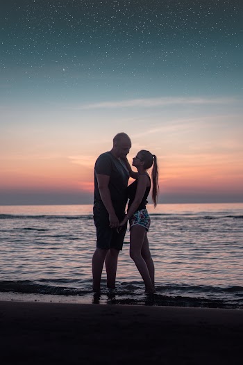 Woman Wearing Black Tank Top and Multicolored Board Short Looking at Man Wearing Black and Gray Crew-neck Shirt and Black Cargo Short Standing on Seashore Sunset Scenery