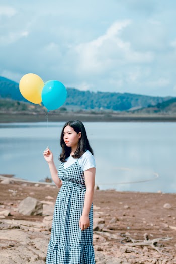 Woman Wearing Blue and White Plaid Dress Holding Blue and Yellow Balloons Near Calm Body of Water at Daytime