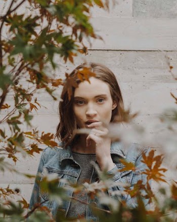 woman wearing blue denim button-up collared jacket holding lip behind brick wall in front of plants