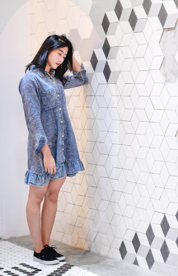 Woman Wearing Blue Denim Button-up Long-sleeved Dress Holding Her Head With Her Left Hand Standing Near White and Gray Concrete Wall