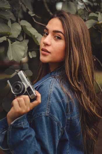 Woman Wearing Blue Denim Jacket While Holding Camera Near Tree
