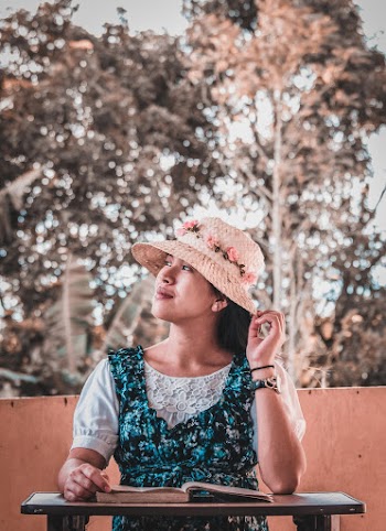 Woman Wearing Blue Floral Dress and Hat Sitting Beside a Table With a Book 