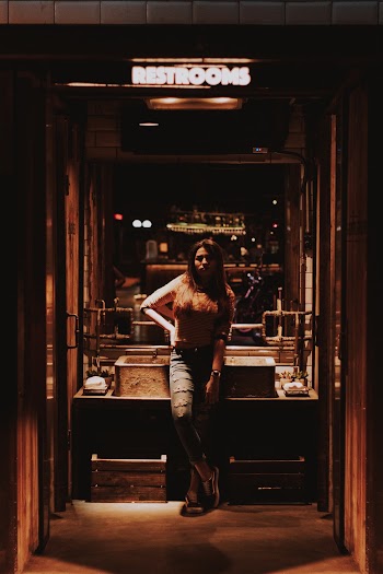 Woman Wearing Denim Jeans Standing Under Restrooms Neon Sign