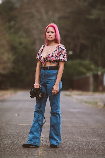 Woman Wearing Floral Crop Top and Blue Denim Jeans Holding Black Dslr Camera Standing in the Middle of Asphalt Road