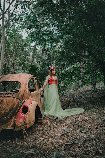 Woman Wearing Green and Red Dress Standing Beside Rusted Brown Volkswagen Beetle Coupe Surrounded by Trees