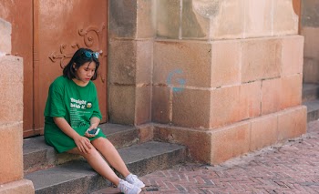 Woman Wearing Green Shirt Sitting Near Brown Gate Holding Smartphone
