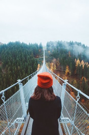 woman wearing knit cap walking on white bridge between trees during daytime