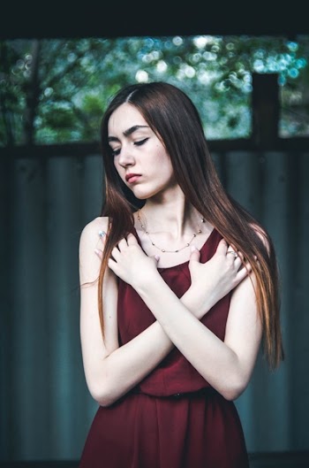 Woman Wearing Maroon Dress With Both Hands on Chest While Closing Eyes Standing Near Wall