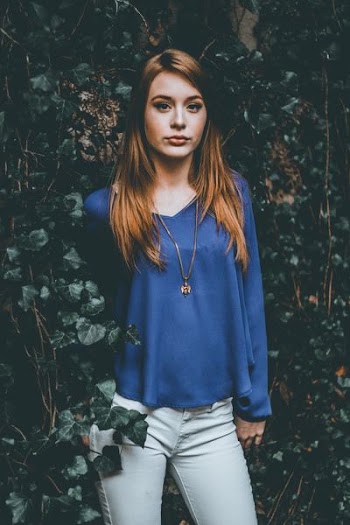 woman wearing necklace, blue top, and white bottoms standing near wall with leaves