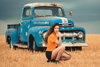 Woman Wearing Orange Shirt And Black Shorts Sitting On Brown Field Near Vintage Blue Pickup Truck