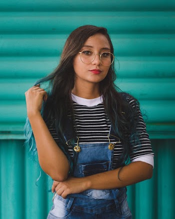 Woman Wearing Overalls And Eyeglasses Standing In Front Of Green Wall