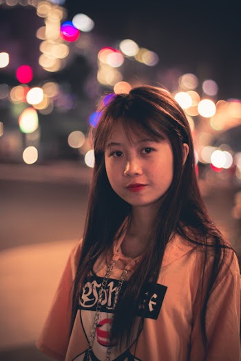 Woman Wearing Pink, Black, and Red Printed Pullover Hoodie Standing Beside Road Bokeh Photography at Night Time