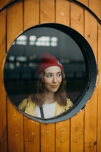 Woman Wearing Red Beanie Standing Near Round Window of Wooden Door