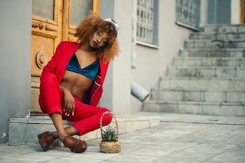 Woman Wearing Red Coat and Pants Sitting Outside Gray Building
