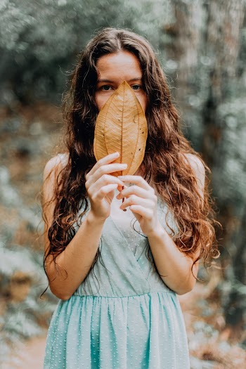 Woman Wearing Teal Dress Holding Brown Leaf In Front Of Her Face