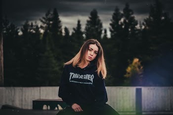 woman wearing Trasher pullover hoodie standing in front of concrete fence and trees