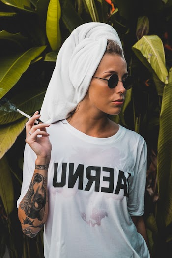 Woman Wearing White Crew-neck Shirt and Black Framed Sunglasses With White Bath Towel on Her Head Holding Cigarette Stick