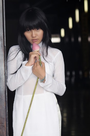 Woman Wearing White Long-sleeved Dress Holding a Pink Lotus Flower Bud Leaning on Grey Wall